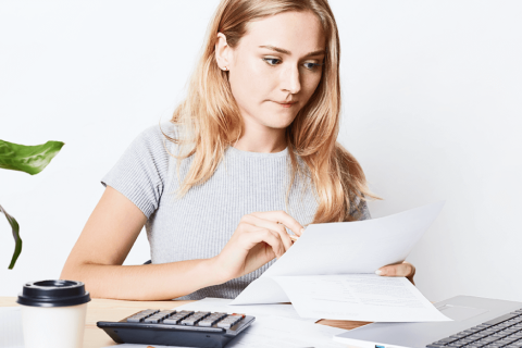 Junge Frau in einem grauen T-Shirt, die konzentriert an einem Schreibtisch sitzt, einen Papierbeleg in der Hand hält und mit einem Laptop arbeitet, während ein Taschenrechner und eine Tasse Kaffee neben ihr liegen.