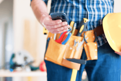 Nahaufnahme eines Bauarbeiters mit Werkzeuggürtel, der auf einer Baustelle sein Smartphone überprüft. Der Bauarbeiter hält einen gelben Schutzhelm und könnte gerade eine Abschlagsrechnung am Handy einsehen oder verwalten.