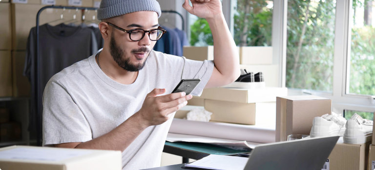Ein junger Unternehmer sitzt inmitten von Kartons und Verpackungsmaterialien vor einem Laptop und freut sich über eine Bestellung auf seinem Smartphone. Er betreibt ein Dropshipping-Geschäft und verwaltet seine Bestellungen online. Im Hintergrund sind Kleidung und Schuhe für den Versand vorbereitet.